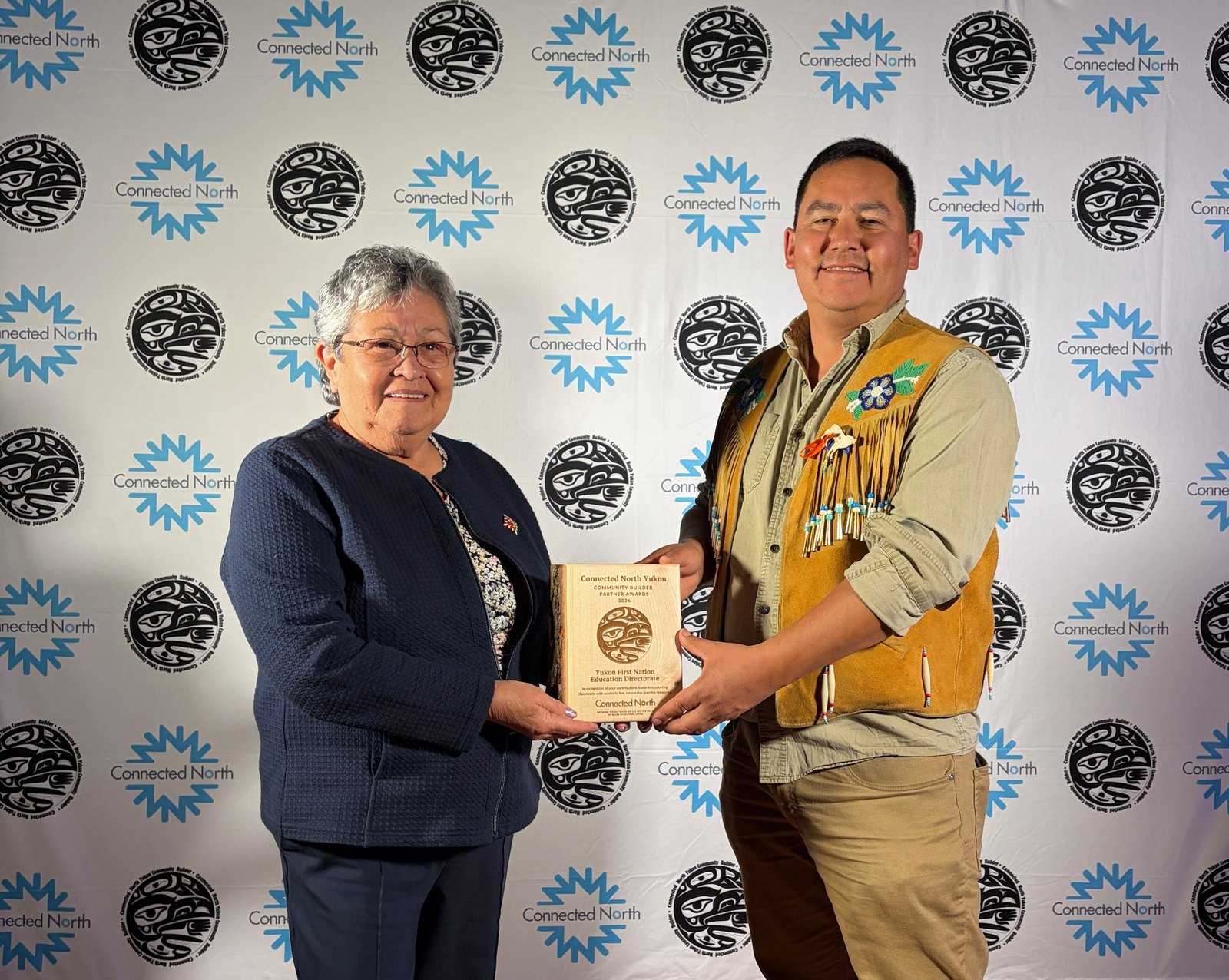 Commissioner Adeline Webber and Chief Sean Uyenets’echᶖa Smith hold a wooden award plaque in front of a Connected North backdrop.
