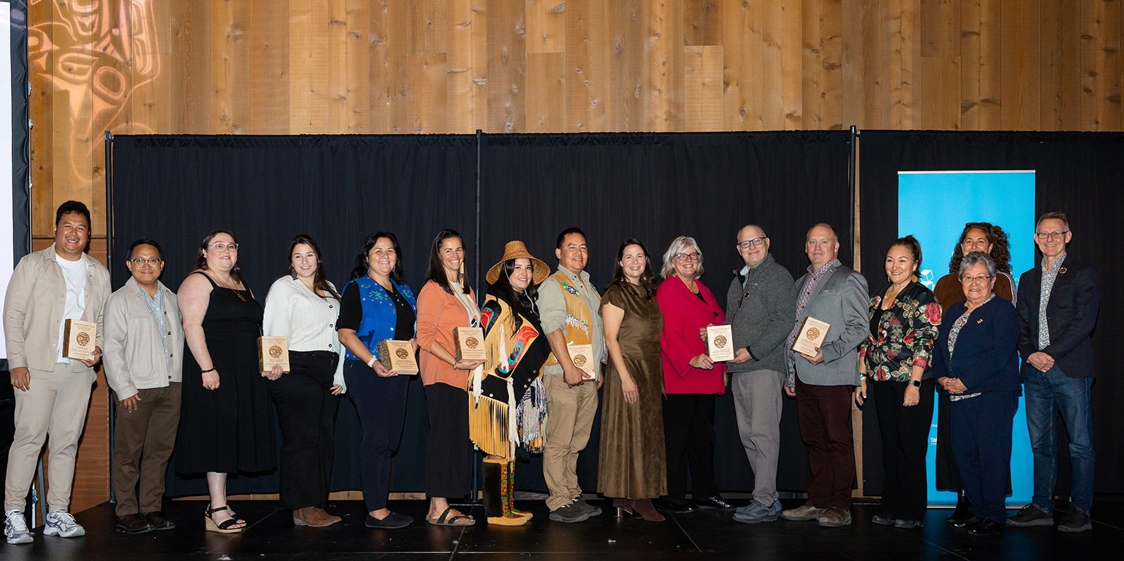A long row of partner representatives and presenters on stage, several holding wooden award plaques.
